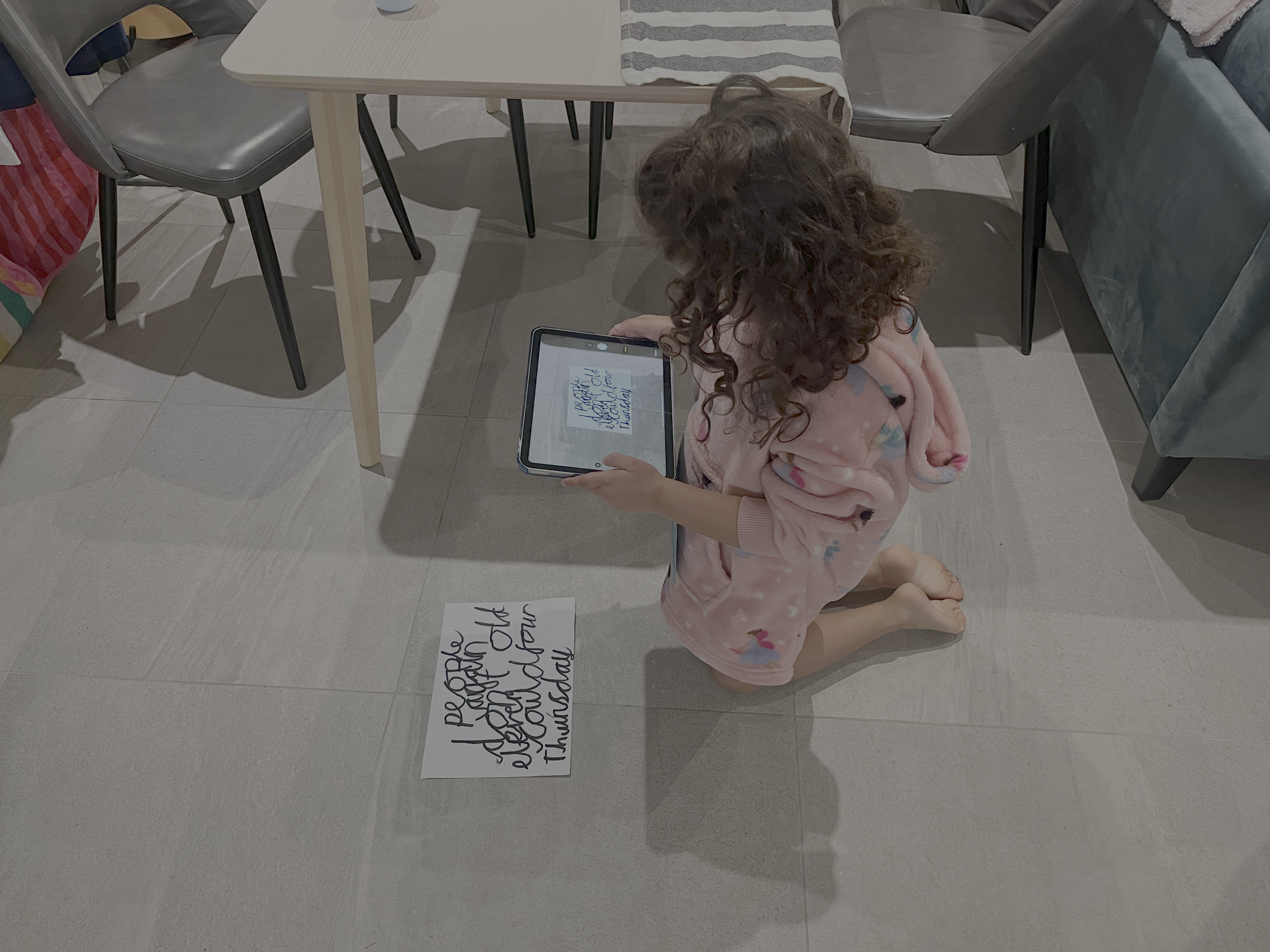 A child sitting on a tiled floor, photographing her handwritten spelling words with a tablet.